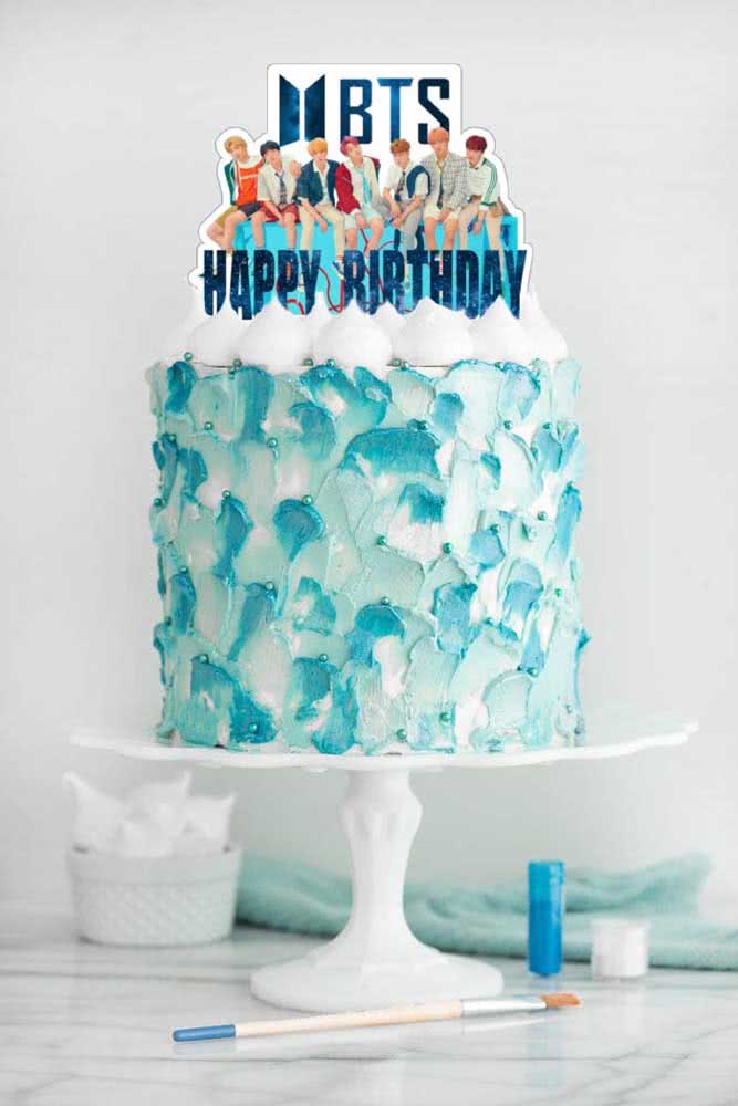 Gâteau BTS décoré de crème fouettée blanche et bleue. Il y a des étiquettes sur le dessus des gâteaux avec des photos de tous les membres.